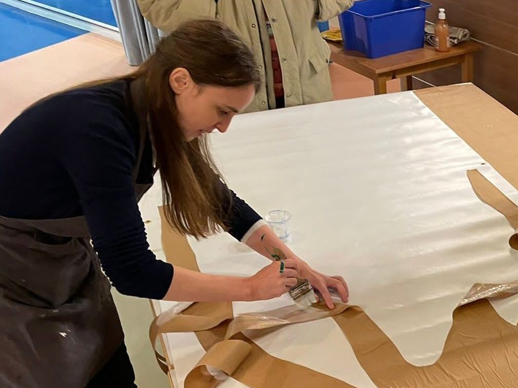 Photo de Sonia Digianantonio en train d'encoller un papier kraft sur un grand papier blanc, en vue d'un atelier d'art plastique.