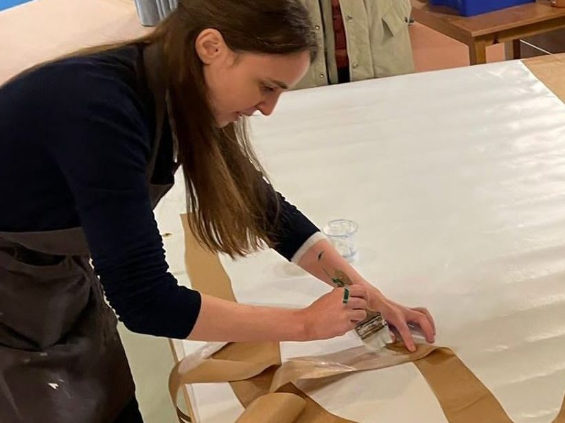 Photo de Sonia Digianantonio en train d'encoller un papier kraft sur un grand papier blanc, en vue d'un atelier d'art plastique.